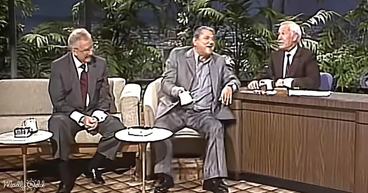 Three men in suits sit on a couch, one laughing, another holding a mug, and the third seated at a desk.