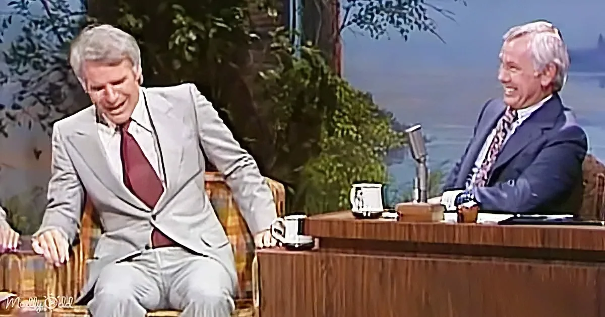 A man in a light suit sits on a couch, laughing, while another man in a dark suit sits behind a desk, smiling.