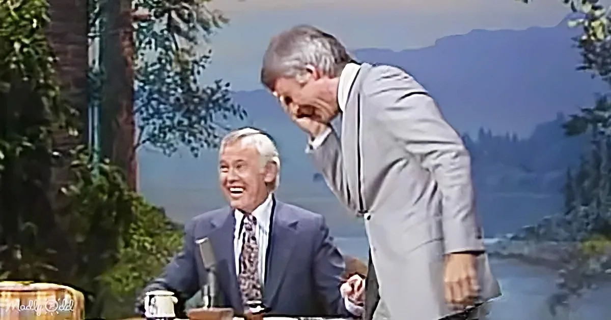 A man in a suit leans over another man in a suit, both smiling, with a scenic backdrop.
