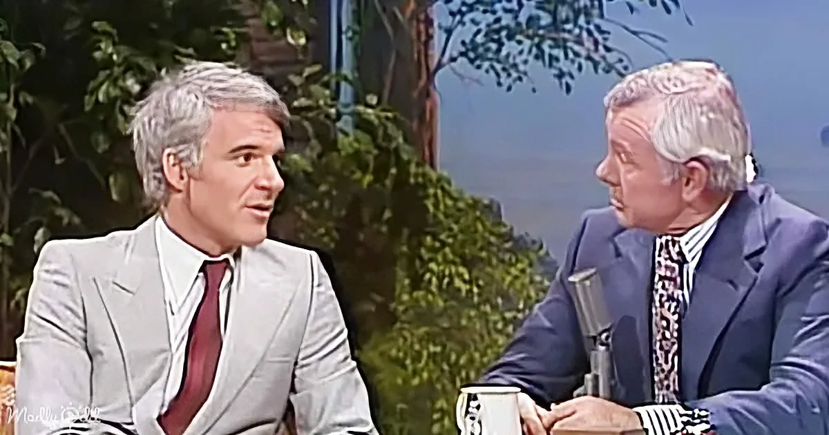 Two men in suits are seated, one in a light gray suit and the other in a dark blue suit, engaged in conversation.