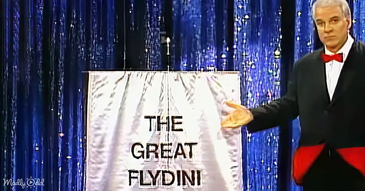 A man in a tuxedo gestures toward a sign reading "The Great Flydini" against a blue curtain backdrop.
