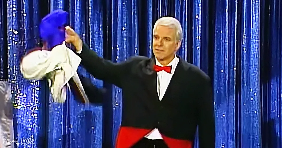 A man in a tuxedo holds a puppet on stage against a blue curtain backdrop.