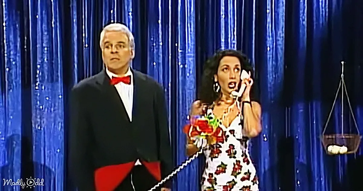 A man in a tuxedo stands beside a woman in a floral dress holding a phone and a bouquet.