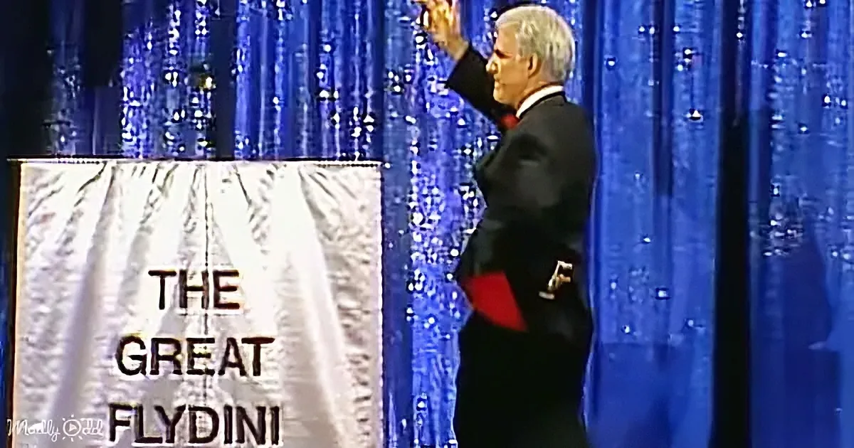 A man in a tuxedo stands on stage, raising his hand, with a banner reading "The Great Flydini" in front of him.