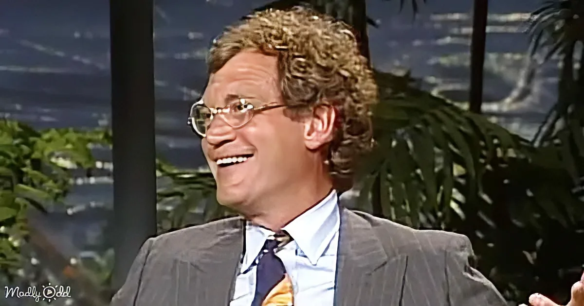 A man with curly hair and glasses smiles in a suit, set against a tropical backdrop.