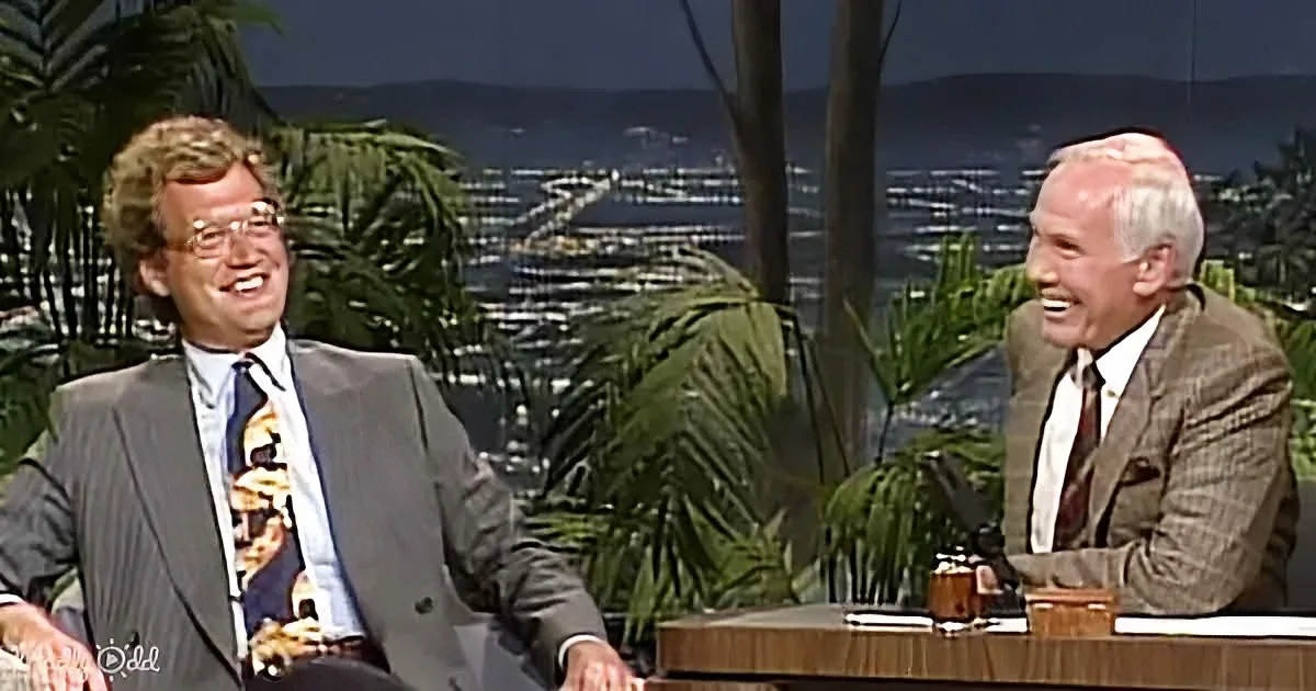 Two men in suits laugh on a talk show set with a cityscape and palm trees in the background.