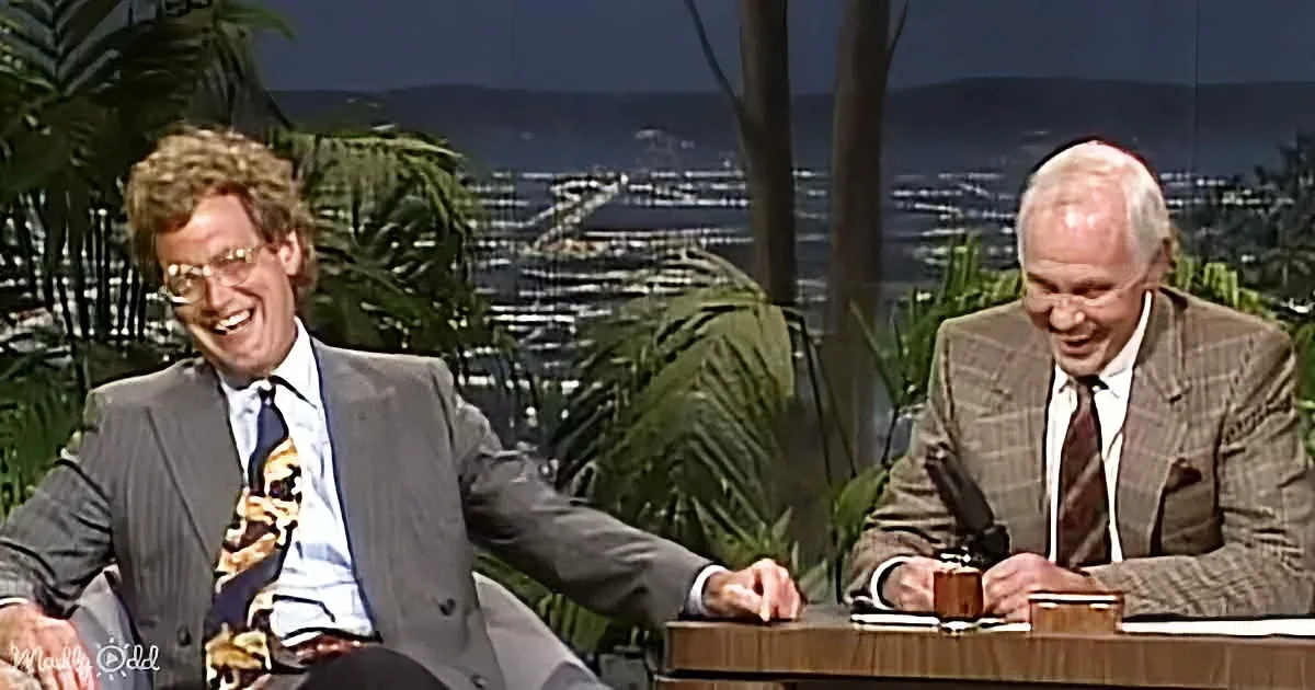 Two men in suits share a laugh on a talk show set with a scenic backdrop.