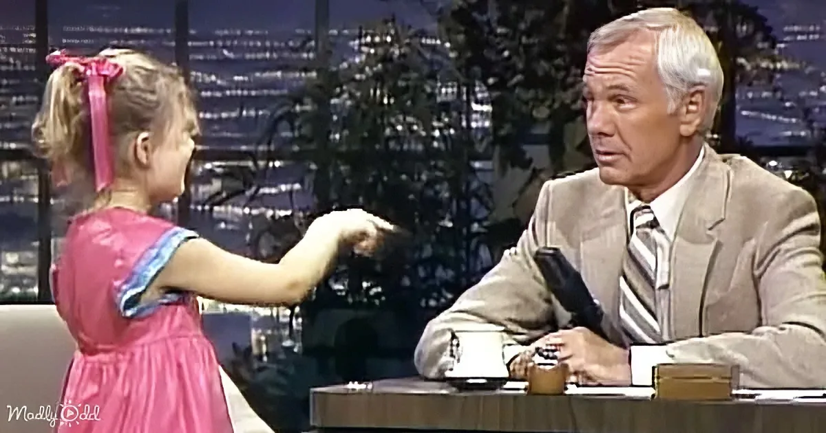 A young girl in a pink dress extends her hand towards a man in a suit on a talk show set.