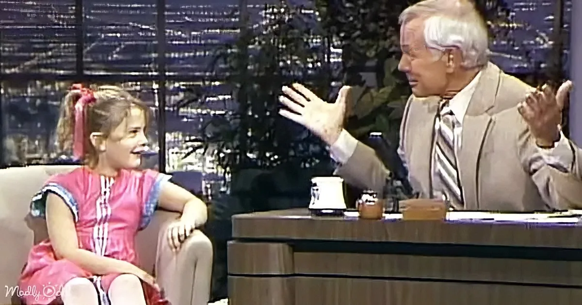 A young girl in a pink dress sits on a couch, smiling, while an older man gestures animatedly during a talk show.