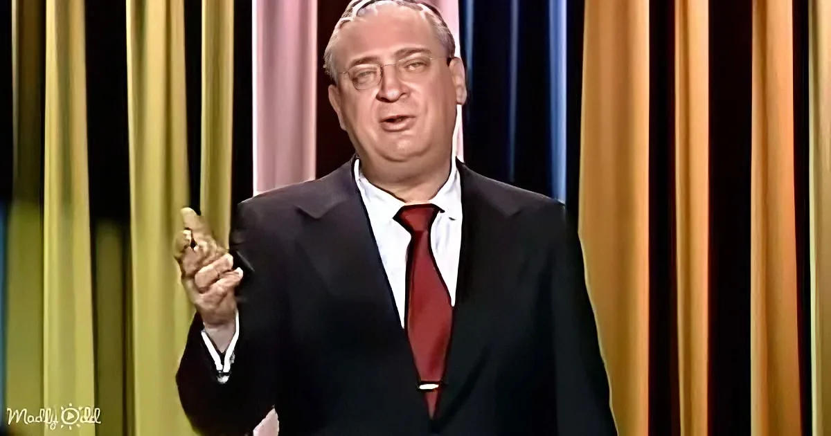 A man in a suit and red tie gestures with his right hand against a colorful striped background.