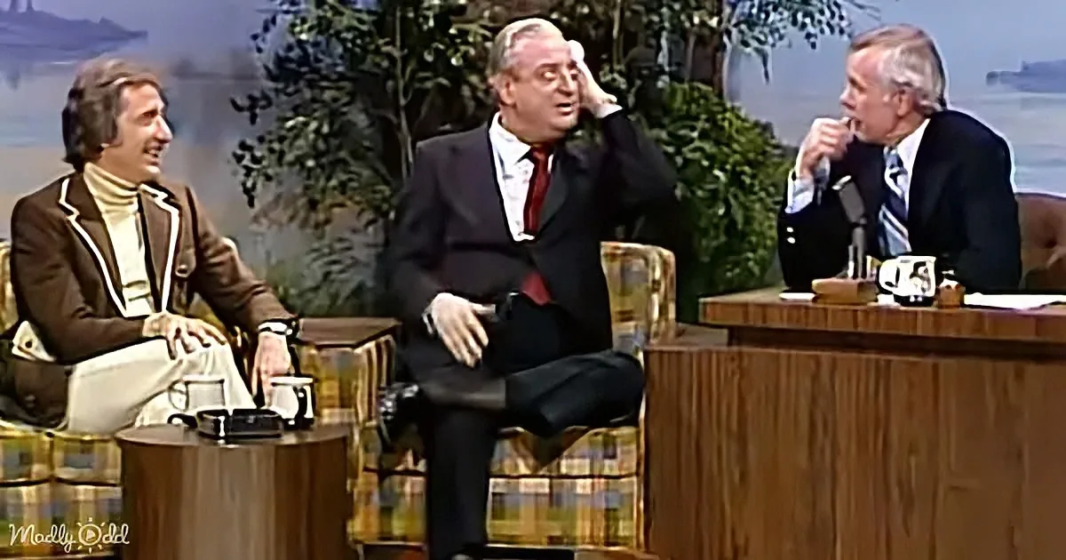 Three men in suits, one gesturing, seated on a talk show set with a backdrop of trees and water.