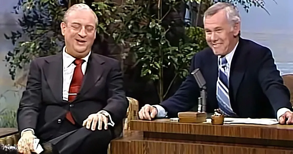 Two men in suits sit at a desk, one smiling, the other laughing, in a studio setting.