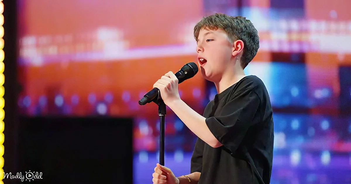 A young boy in a black shirt sings into a microphone on a stage with colorful lights.