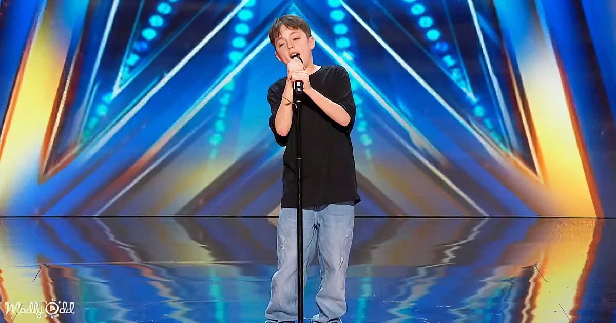 A young boy stands on stage, singing into a microphone, wearing a black shirt and light blue jeans.