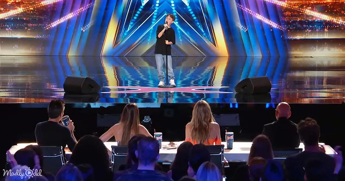 A young person sings on stage with a microphone, judges sit at a table in front of them.
