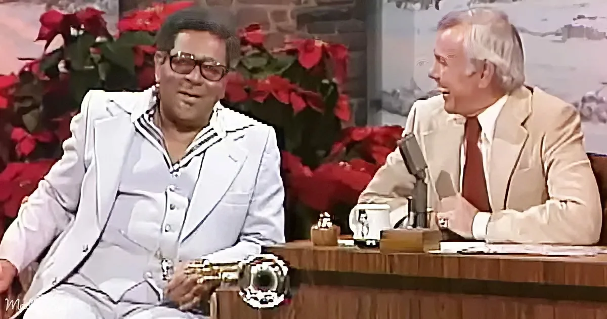 A man in a white suit sits on a couch, smiling, while another man in a beige suit sits at a desk with a microphone, in...