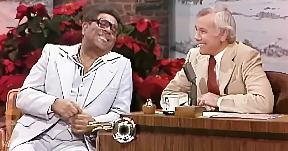 Two men in suits, one in white, laughing on a talk show set with a festive background.