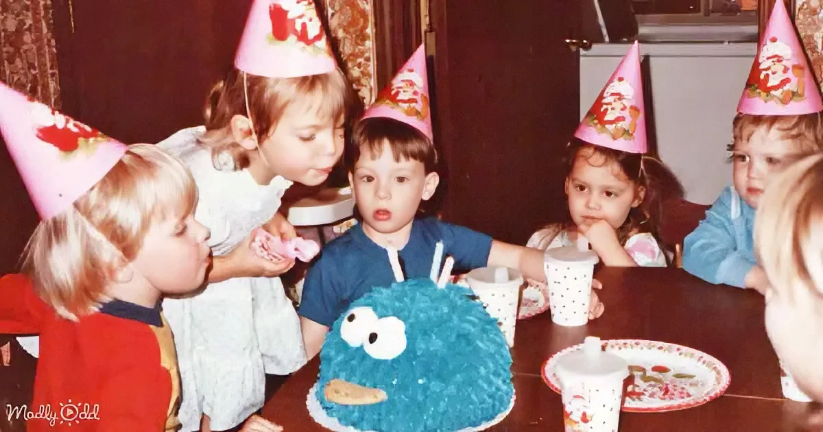 Children wearing party hats gather around a table with a Cookie Monster cake and cups.