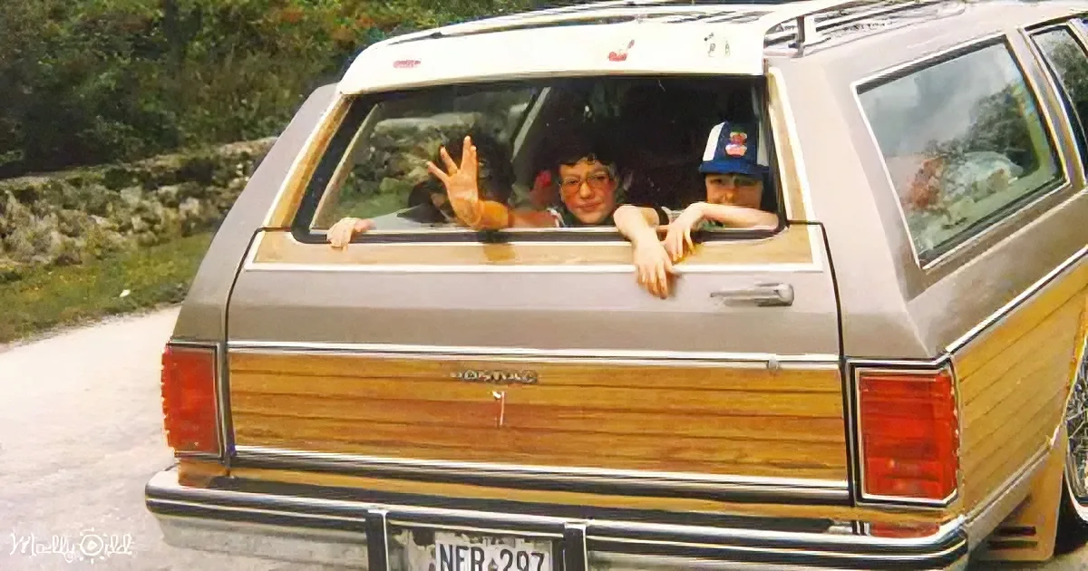 A vintage station wagon with wood paneling has two people waving from the back window.