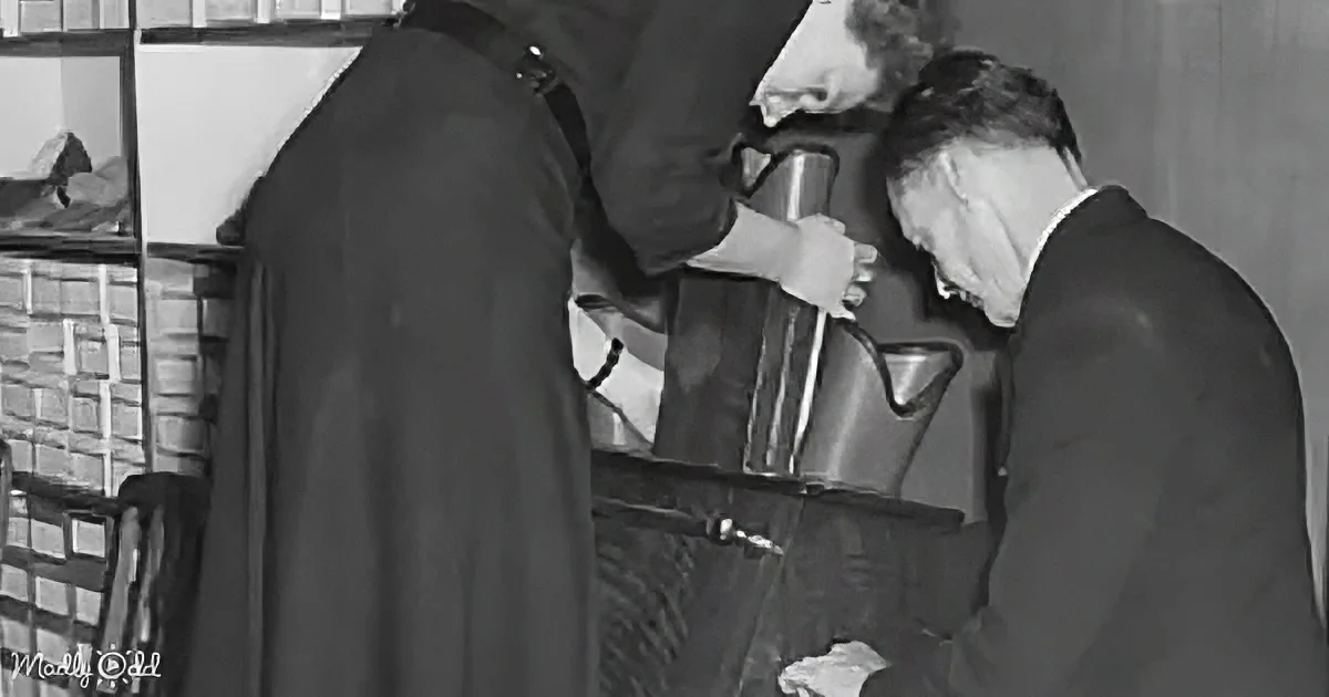 A man in a suit pours water from a metal pitcher into a glass, while another person in a cloak leans in.