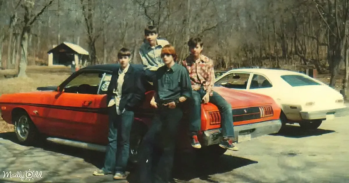 Four young men pose with a red vintage car in a wooded area.
