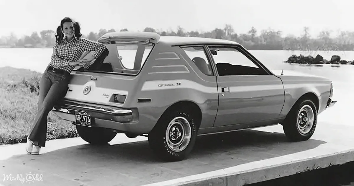 A woman leans against a vintage AMC Gremlin X parked near a body of water.
