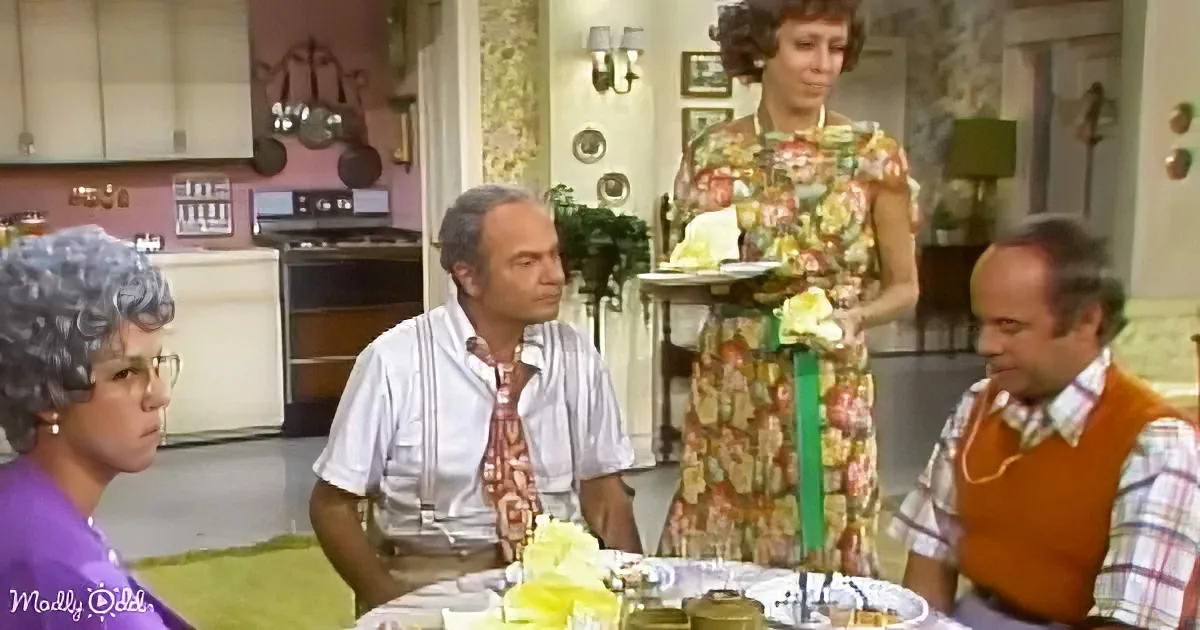 Woman in floral dress holds tray, stands by table with seated individuals in retro kitchen.