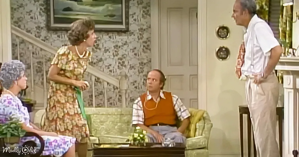 Woman in floral dress stands, gesturing, while man in orange vest sits on couch, and another man stands nearby in a...