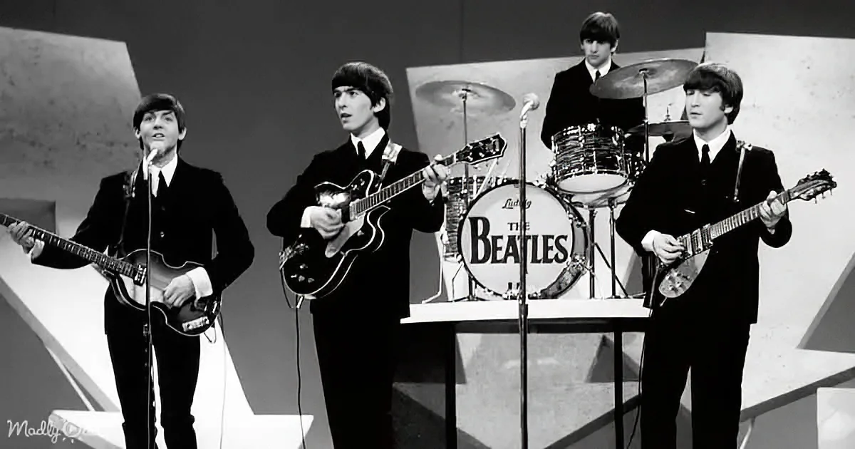 The Beatles perform on stage, dressed in black suits, with George Harrison playing the guitar and Ringo Starr behind the...