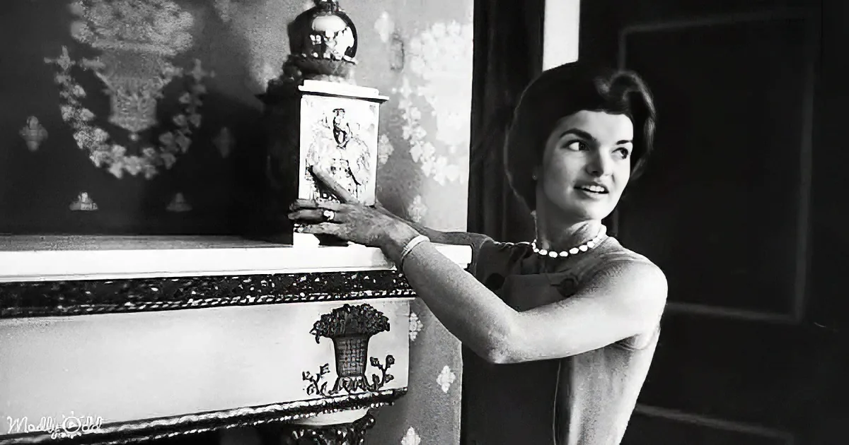 A woman in a polka-dot dress and pearl necklace leans on a decorative dresser, smiling.