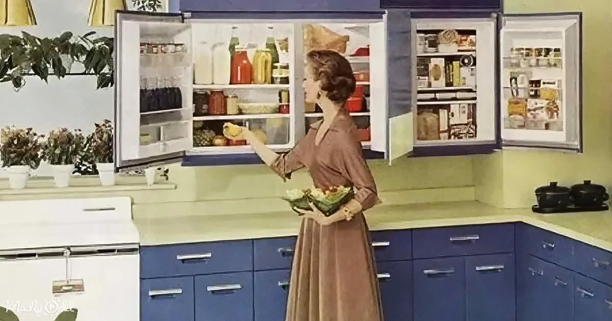 A woman in a brown dress holds a salad bowl, reaching into a large, open refrigerator in a retro kitchen.