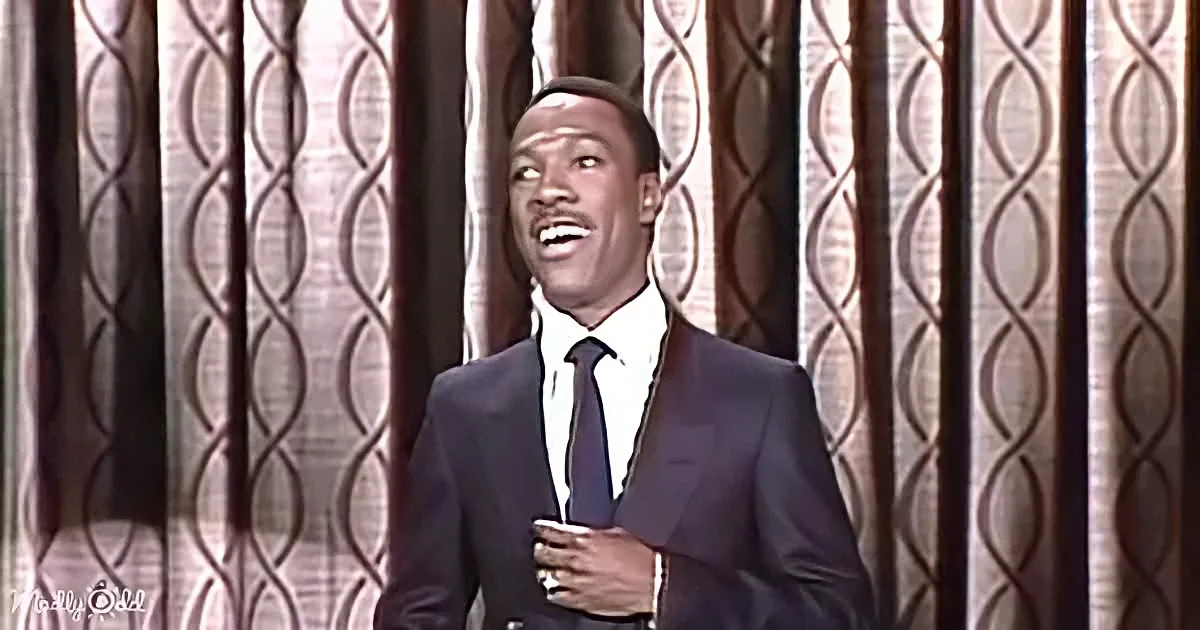 A man in a suit and tie is smiling and holding a microphone, standing in front of patterned curtains.