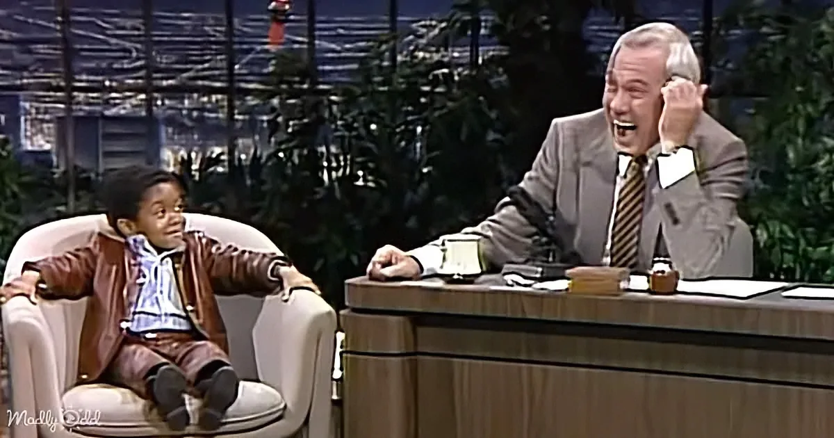 A young boy in a brown jacket sits in a chair, while a man in a suit laughs on a talk show set.