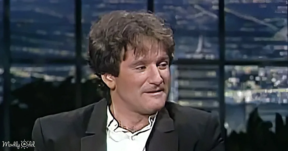 A man with curly hair, wearing a black suit and white shirt, is seated in front of a cityscape backdrop.