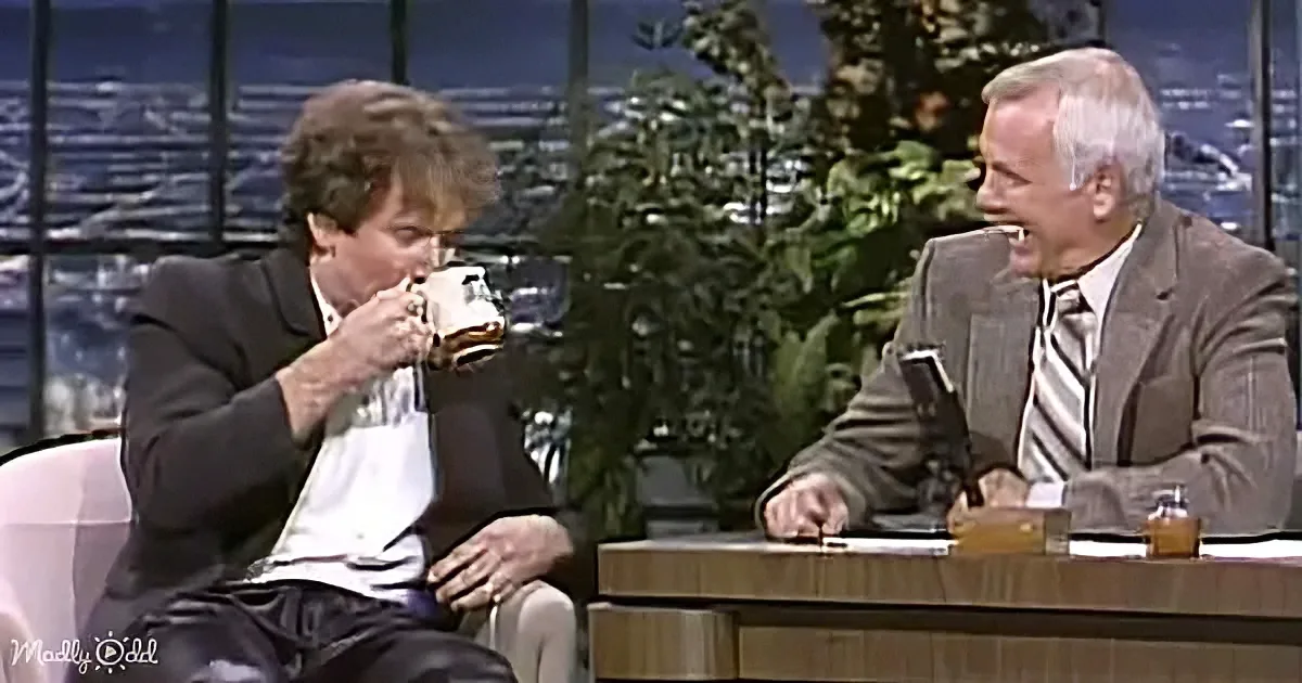 A man drinks from a mug while another sits at a desk, both in a talk show setting.