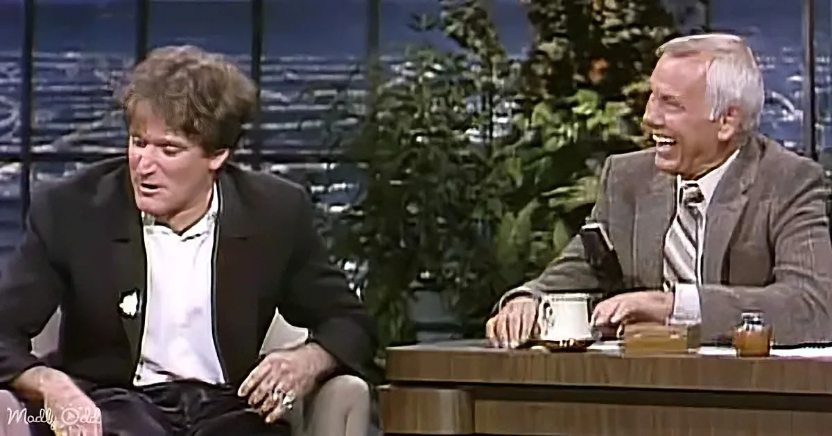 A man in a suit sits on a talk show set, laughing with another man in a suit.
