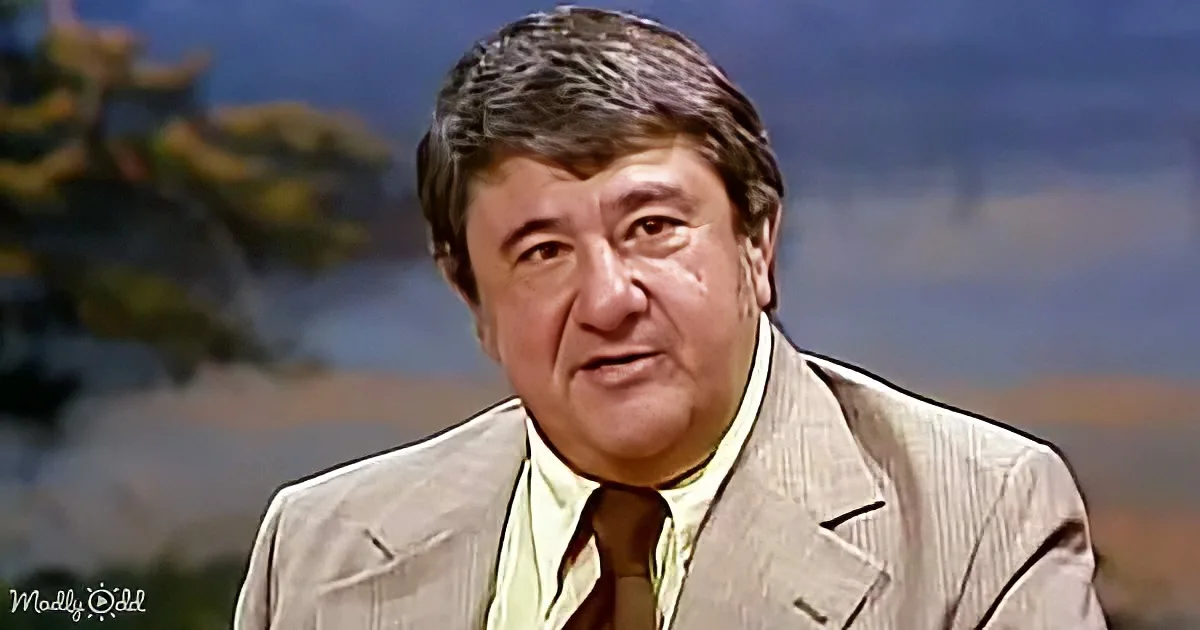 A man in a beige suit and green shirt is speaking, with a blurred background of trees and sky.