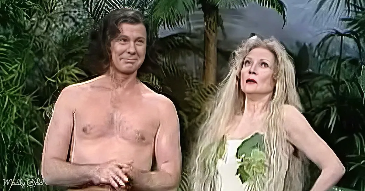 A man with a hairy chest and a woman in a green dress stand in a jungle setting.