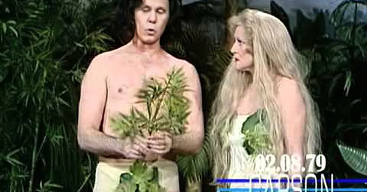 A man in a pink outfit with arms crossed stands next to a woman in a white dress with green leaves, both smiling in front...