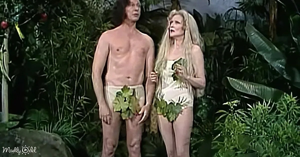 A man and woman stand in a jungle setting, both holding leaves, with the woman appearing surprised.