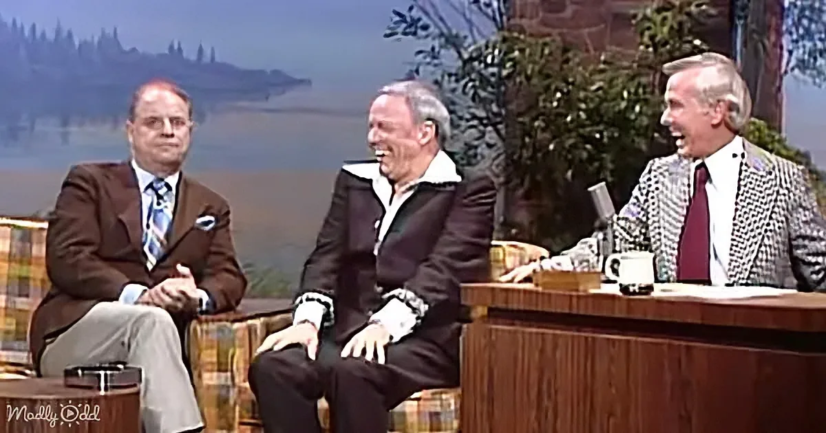 Three men in suits sit on a talk show set, laughing and engaging in conversation.