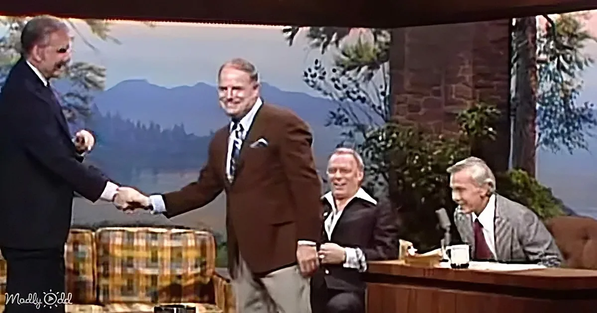 Two men in suits shake hands on a talk show set with a scenic backdrop.