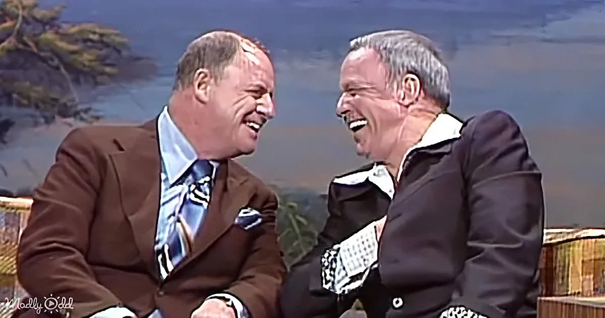 Two men in suits share a laugh on a talk show set with a scenic backdrop.