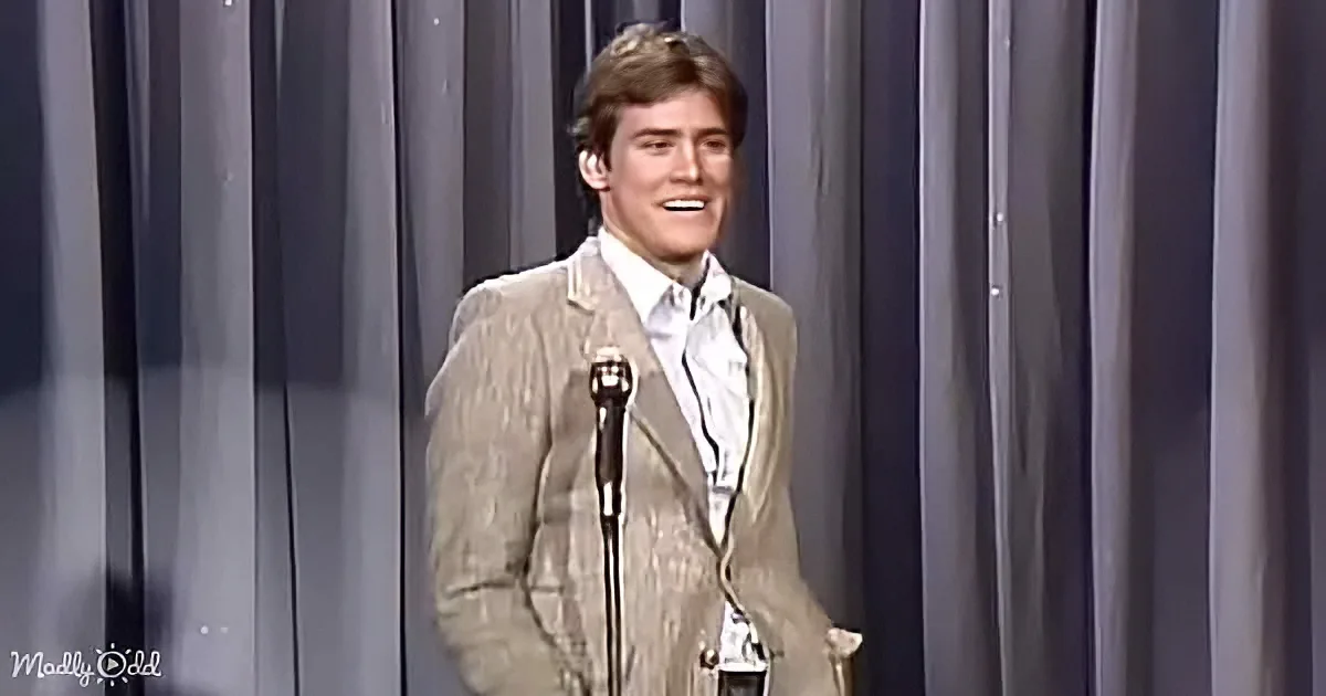 A man in a beige suit stands on stage with a microphone, smiling, against a gray curtain backdrop.