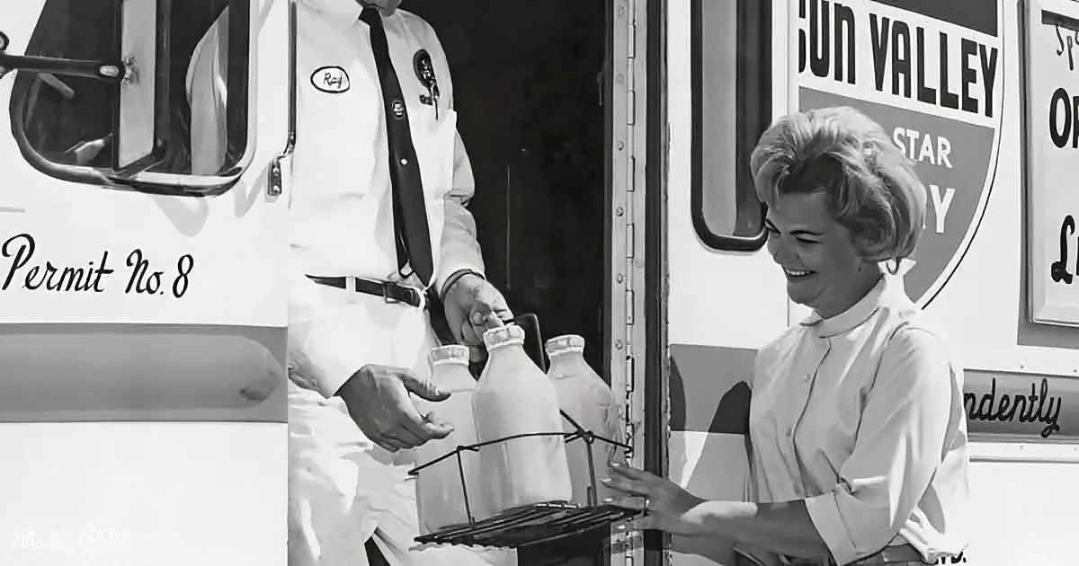 A milkman in uniform delivers milk to a smiling woman in front of a delivery truck.