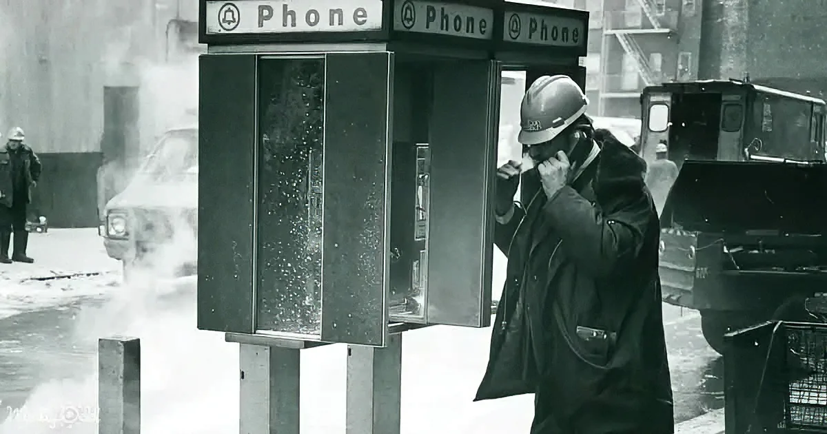 A man in a hard hat talks on a payphone in a snowy urban setting.