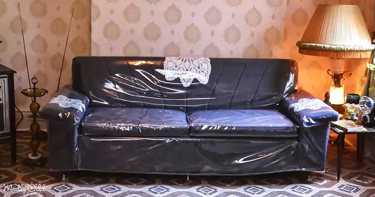A black couch is covered in plastic wrap, placed against a patterned wall with a lamp and table nearby.