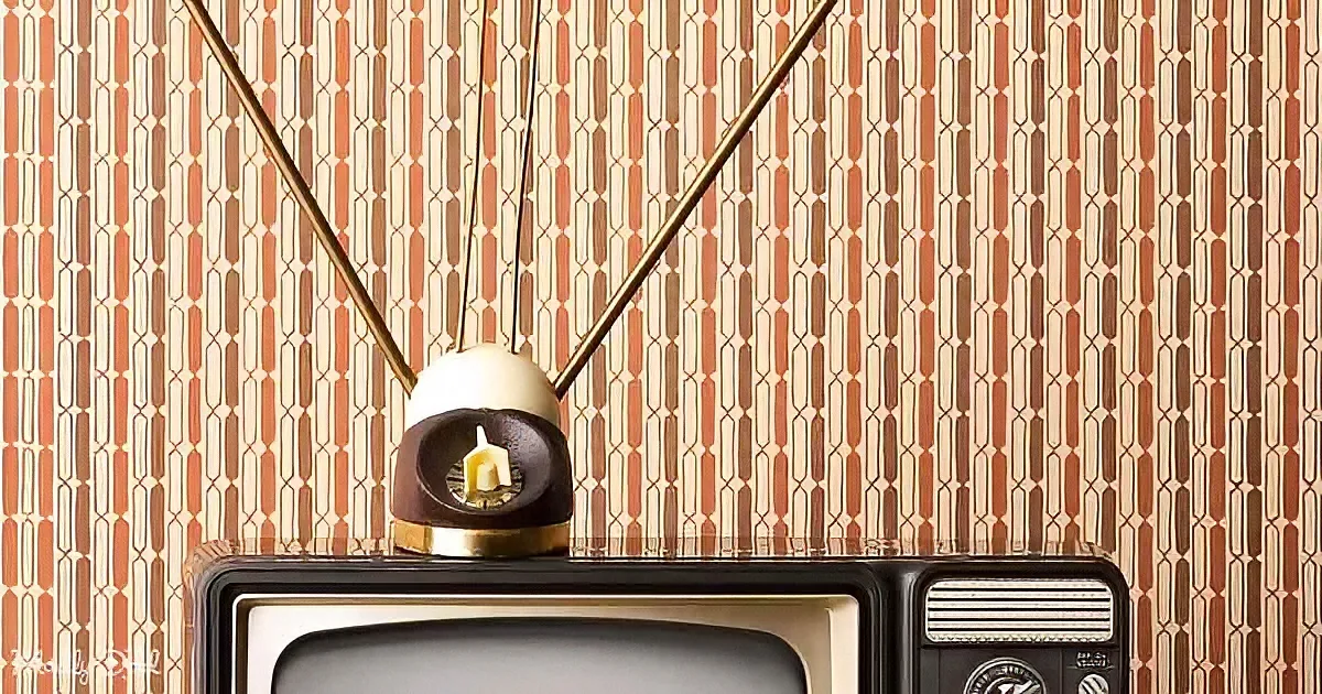 A vintage TV with a unique antenna sits atop a retro TV, against a patterned wallpaper backdrop.