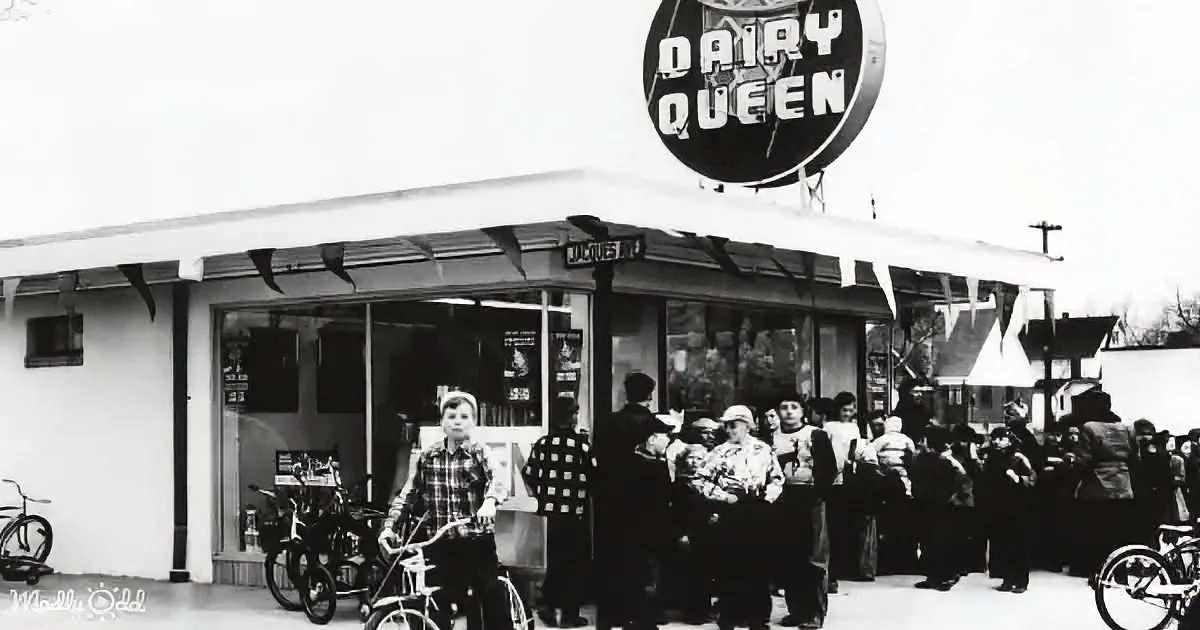A young boy on a bicycle stands in front of a Dairy Queen, with people waiting in line and bicycles parked nearby.
