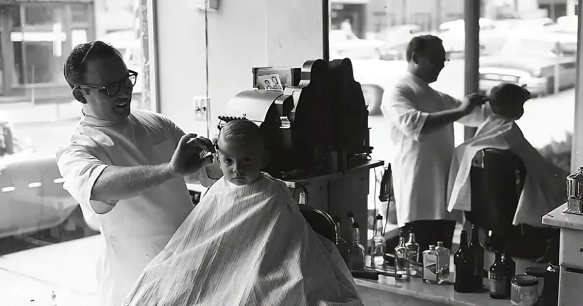 A barber in a white shirt cuts a child's hair in a barbershop with another barber in the background.